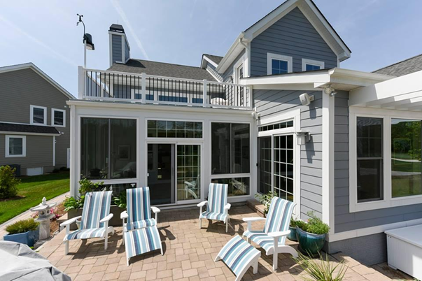 The exterior of a beach-style home with a paved patio attached to it with outdoor chairs and a one-story patio room on the side of the house with an open deck above it.